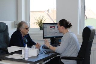 Eine Ärztin berät eine Seniorin am Schreibtisch, am Computer ist ein Diagramm zu sehen.